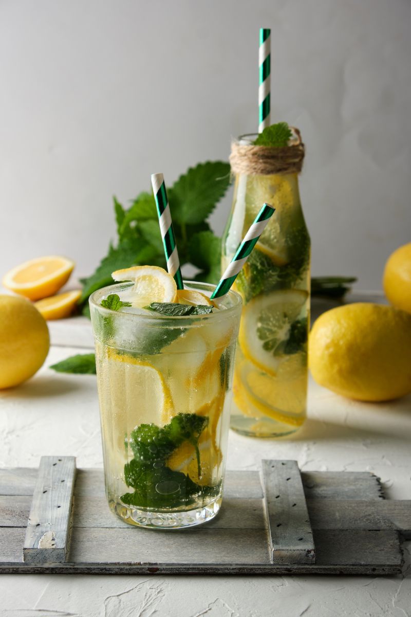 Tradycyjna lemoniada z plasterkami cytryny i świeżą miętą, podana w wysokiej szklance oraz w szklanej butelce, ustawionej na białym stole w naturalnym świetle.