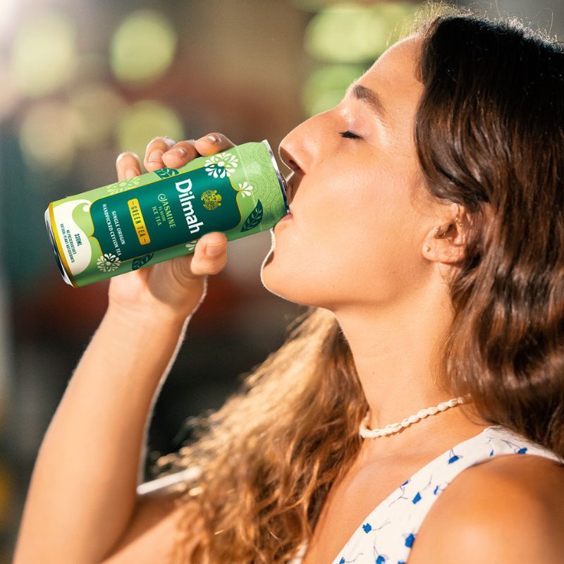 Kobieta delektująca się orzeźwiającą herbatą Dilmah Jasmine Flavor Green Ice Tea w letniej atmosferze. W jej mimice widać czystą przyjemność. Zdjęcie emanuje lekkością i beztroską typową dla ciepłych, słonecznych dni.