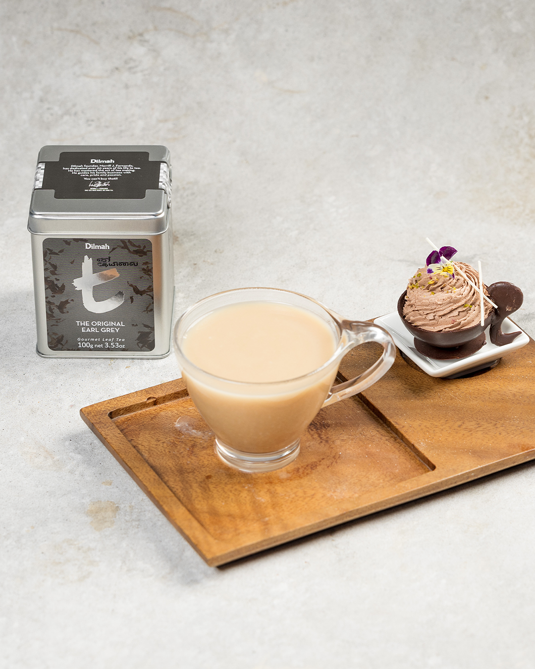 Herbata Earl Grey z mlekiem podana w przezroczystym kubku. Obok deser czekoladowy i opakowanie Dilmah Original Earl Grey.