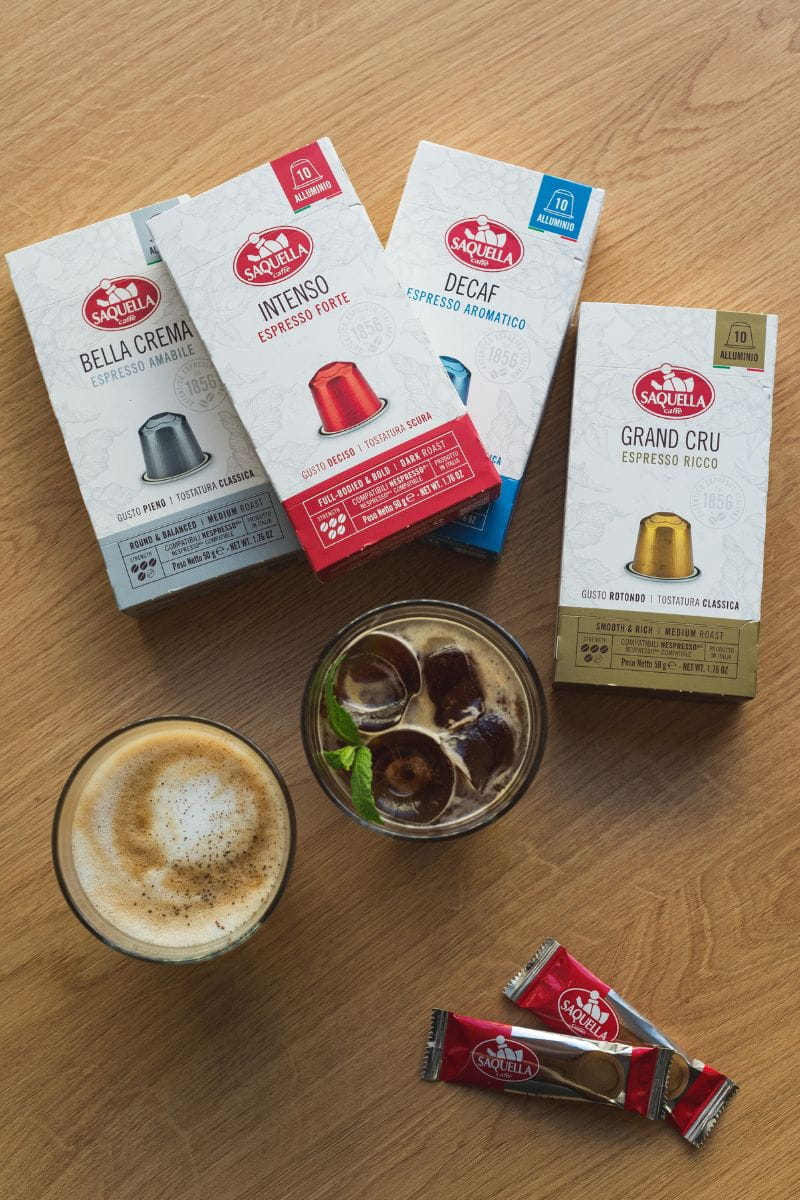 Na stole, obok dwóch szklanek z kawą i cukru w saszetkach leżą opakowania kawy w kapsułkach Saquella – od lewej Saquella Bella Crema Espresso Amabile, Saquella Intenso Espresso Forte, Saquella Decaf Espresso Aromatico I Saquella Gran Cru Espresso Ricco.