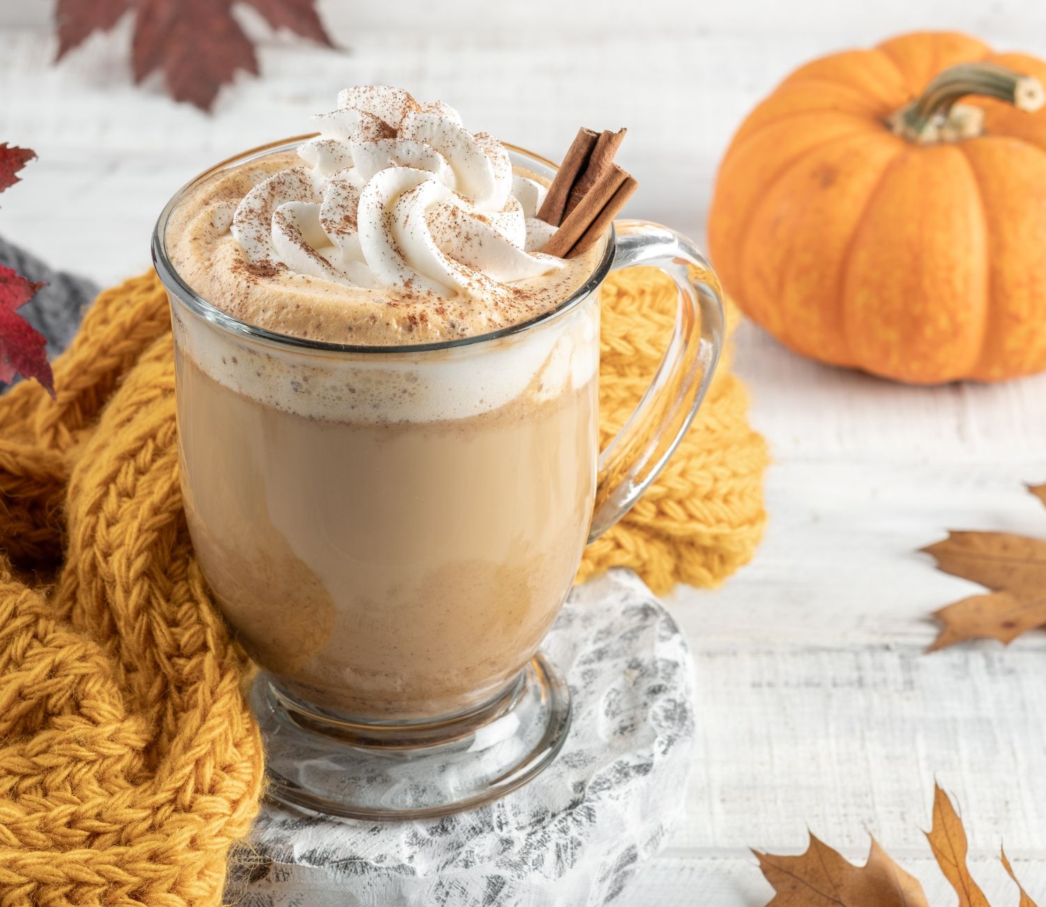 Pumpkin Spice Latte w przezroczystym kubku, ozdobione bitą śmietaną i posypane cynamonem. W tle jesienne dekoracje, szalik, kolorowe liście i dynia.