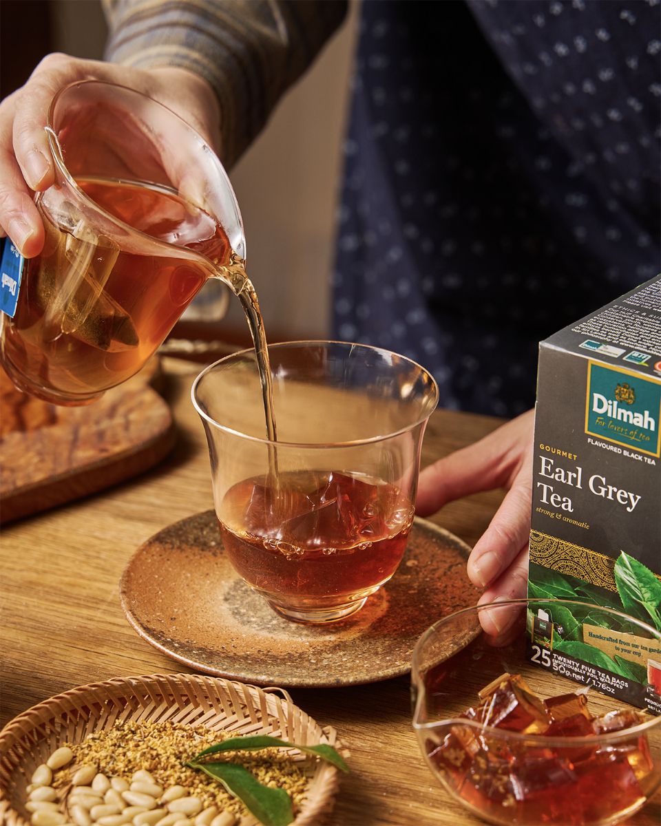 Mężczyzna wlewa herbatę Dilmah Earl Grey Tea do szklanki. Na stole stoi opakowanie z herbatą Earl Grey, kostki lodu, taca z przyprawami i owoc bergamotki.
